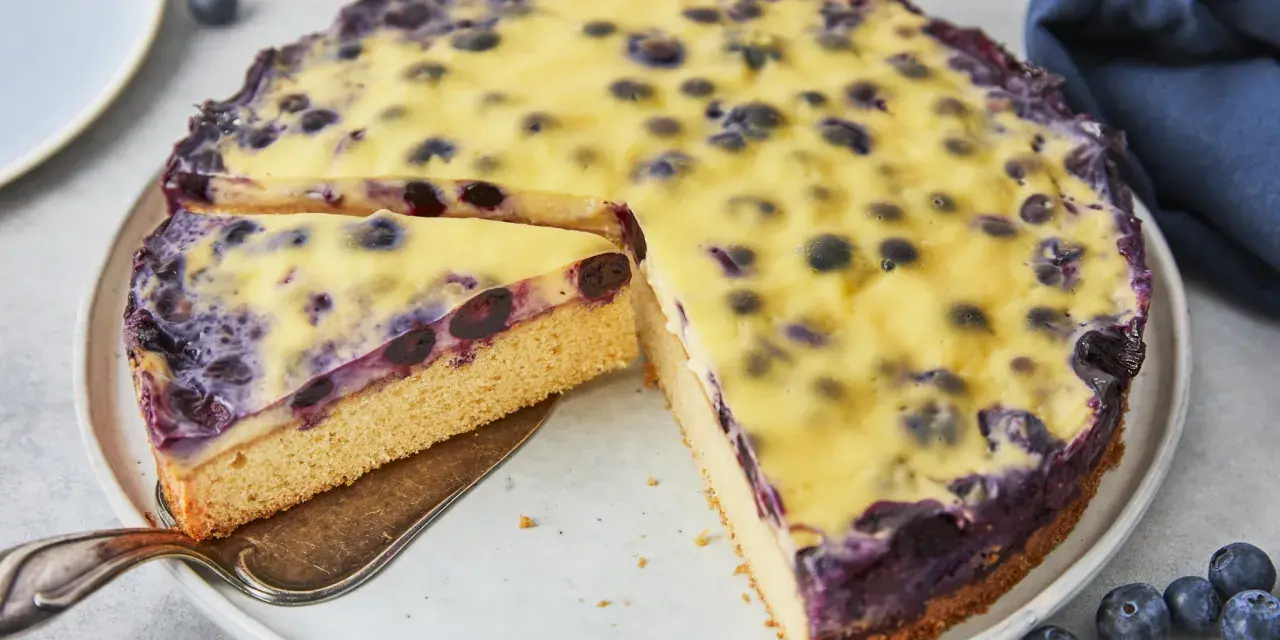 Ein saftiger Heidelbeerkuchen mit einer cremigen Vanillepudding-Schicht und frischen Heidelbeeren. Ein Stück wird gerade herausgeschnitten.