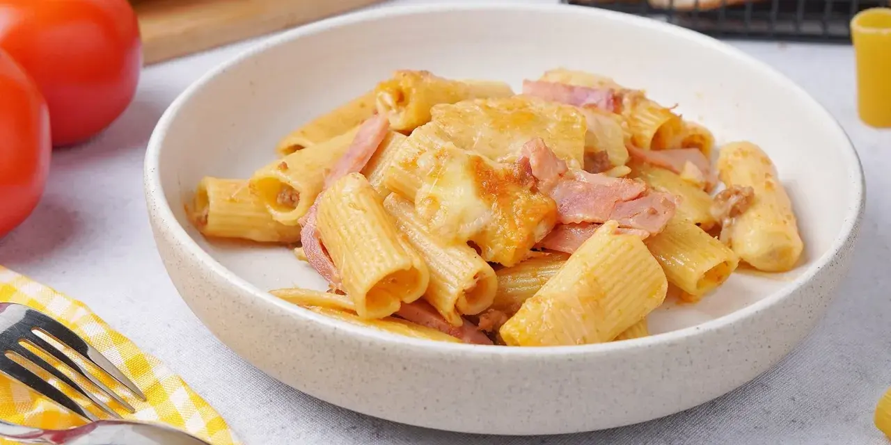 Herzhaftes Rigatoni al Forno mit Schinken und geschmolzenem Käse in einer Schüssel, daneben Tomaten und Brot.