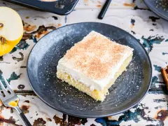 Ein Stück von Omas Apfelmuskuchen vom Blech, cremig belegt und mit Zimt bestreut, daneben ein halber Apfel und Zimtstangen.
