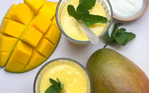 Zwei Gläser cremiger Mango Lassi, garniert mit Minze, neben frischen Mangos und einem Schälchen Joghurt.