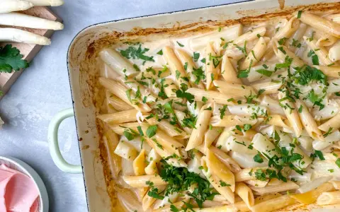 Cremige Spargel Pasta mit frischer Petersilie in einer Auflaufform. Dazu liegen Spargelstangen, Schinken und Parmesan bereit.