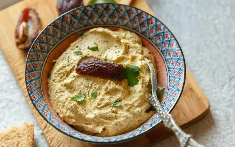 Cremiger Dattel Dip in einer bunten Schale, garniert mit einer Dattel und Petersilie. Dazu Brot und weitere Datteln auf einem Holzbrett.