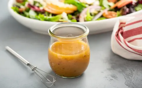 Ein Glas mit honig senf dressing steht neben einem kleinen Schneebesen. Im Hintergrund eine Schüssel mit Salat.