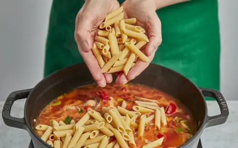 Hände geben Penne in einen Topf mit köchelnder Sauce für ein einfaches One-Pot-Chicken-Pasta-Gericht.