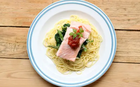Ein Teller mit One Pot Lachs Pasta, garniert mit Petersilie und Tomatensauce, daneben Spargel und Spinat auf einem Holztisch.