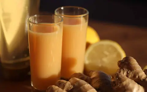 Zwei Gläser mit einem leuchtend orangen Ingwer Shot stehen neben frischem Ingwer und Zitronenscheiben.