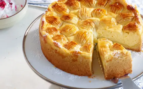 Ein köstlicher, schneller Apfelkuchen mit Schmand und Pudding, verziert mit Apfelscheiben und Puderzucker. Daneben eine Schüssel mit Beeren-Sahne.