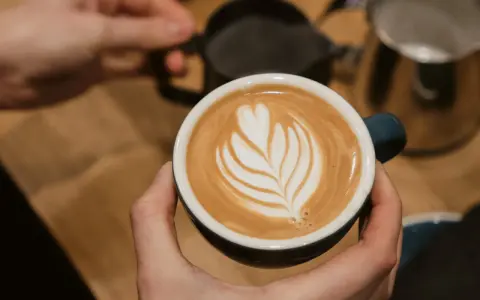 Cappuccino selber machen: Hände halten eine Tasse mit Latte Art. Im Hintergrund Milchkännchen und Sieb.