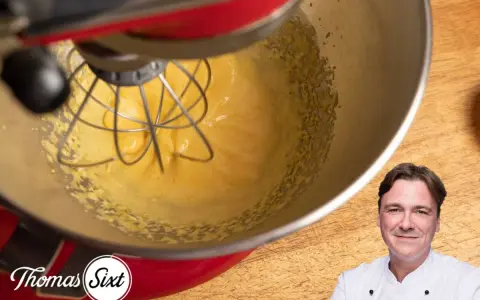 Küchenmaschine mixt eine cremige, gelbe Masse, die an Sauce Hollandaise erinnert. Ein Koch lächelt im Vordergrund.