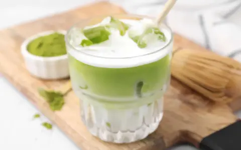 Erfrischender Matcha Latte mit Eis, Milch und einem Hauch von Starbucks-Rezept-Magie. Daneben eine Schale Matcha-Pulver und ein Chasen.