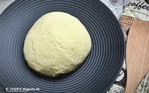 Ein runder Pizzateig auf schwarzer Platte wartet darauf, zu wissen, wie lange muss ein pizzateig ruhen, bevor er perfekt wird.