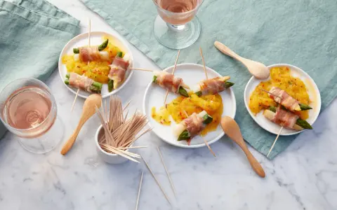 Leckere Spargel im Speckmantel auf gelbem Chutney, serviert auf kleinen Tellern mit Holzspießen und Roséwein.