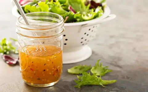 Ein Glas mit selbstgemachtem Salatdressing, das aussieht wie im Restaurant. Daneben ein Sieb mit frischem Salat.