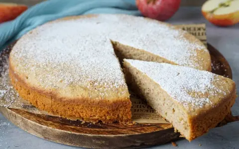 Ein schneller, saftiger Kuchen mit Apfelmus, bestäubt mit Puderzucker. Ein Stück wird gerade herausgenommen.
