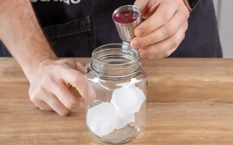 Eiswürfel in einem Glas, dazu wird rote Flüssigkeit aus einem Messbecher gegossen. So einfach lässt sich Rhabarberschorle selber machen!