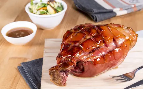 Glänzende, karamellisierte Schweinshaxe sous vide auf Holzbrett, daneben Schüssel mit Sauce und Salat.