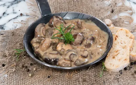 Herzhafte Champignon Rahmsauce in einer Pfanne, garniert mit Rosmarin, daneben knuspriges Brot und Pfefferkörner.