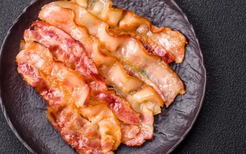 Knusprig gebratener Bacon im Ofen, auf einem dunklen Teller angerichtet.