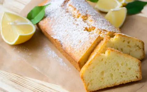 Saftiger veganer Zitronenkuchen, bestäubt mit Puderzucker, mit frischen Zitronenspalten und Blättern garniert.