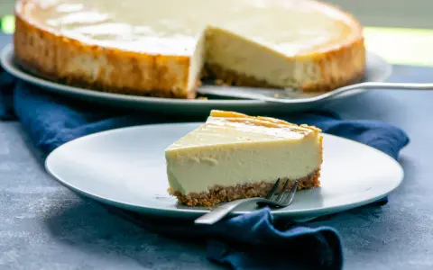 Ein Stück cremiger american cheesecake mit knusprigem Boden auf einem Teller, daneben ein ganzer Kuchen.