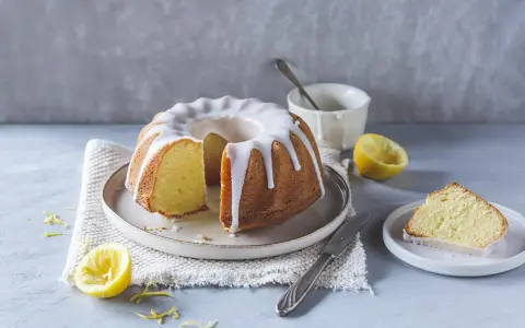 Ein saftiger Zitronenkuchen mit Zuckerguss, perfekt für ein Lemon Drizzle Cake Rezept. Daneben liegen frische Zitronen und ein Stück Kuchen.