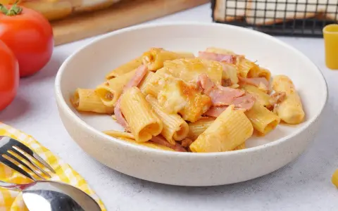 Herzhaftes Rigatoni al Forno mit Schinken und geschmolzenem Käse in einer Schüssel, daneben Tomaten und Brot.