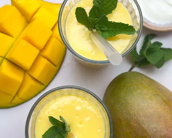 Zwei Gläser cremiger Mango Lassi, garniert mit Minze, neben frischen Mangos und einem Schälchen Joghurt.