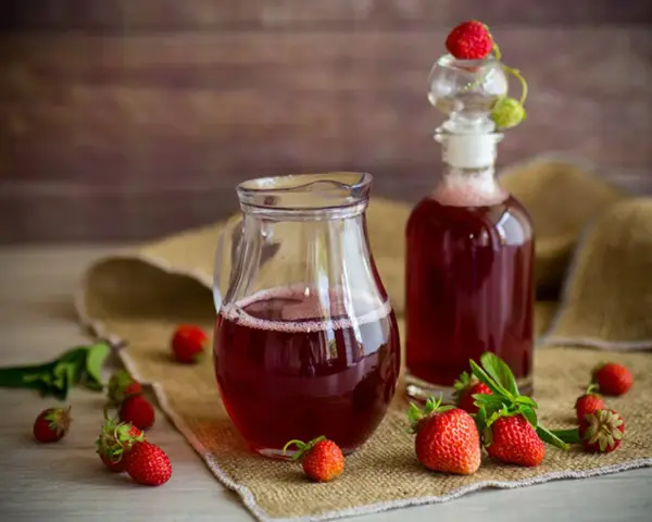 Frischer Erdbeersirup in Karaffe und Flasche, umgeben von reifen Erdbeeren. Perfekt, um Erdbeersirup selber machen.