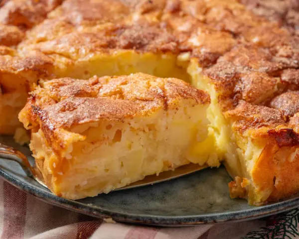 Ein saftiger apfelkuchen mit schale, goldbraun gebacken und mit Zimt bestreut. Ein Stück wird gerade herausgehoben.