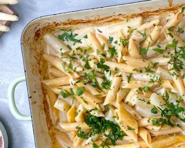 Cremige Spargel Pasta mit frischer Petersilie in einer Auflaufform. Dazu liegen Spargelstangen, Schinken und Parmesan bereit.