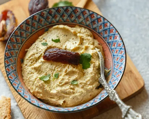 Cremiger Dattel Dip in einer bunten Schale, garniert mit einer Dattel und Petersilie. Dazu Brot und weitere Datteln auf einem Holzbrett.