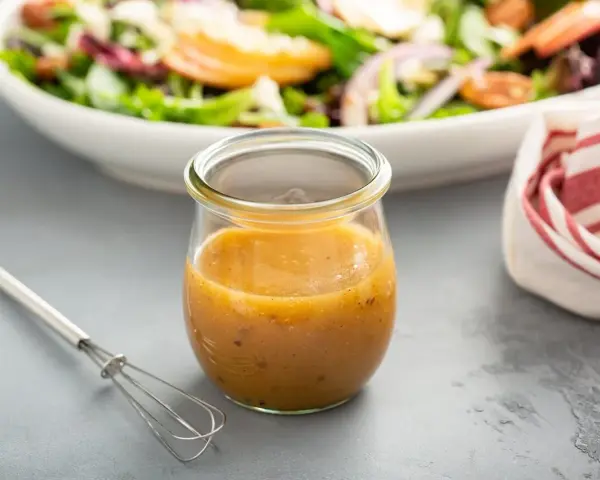 Ein Glas mit honig senf dressing steht neben einem kleinen Schneebesen. Im Hintergrund eine Schüssel mit Salat.
