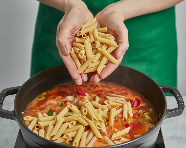 Hände geben Penne in einen Topf mit köchelnder Sauce für ein einfaches One-Pot-Chicken-Pasta-Gericht.