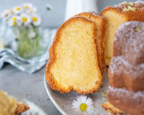 Saftiger Omas Joghurt Kuchen, frisch gebacken und mit Puderzucker bestäubt, daneben eine Zitronenscheibe und Gänseblümchen.