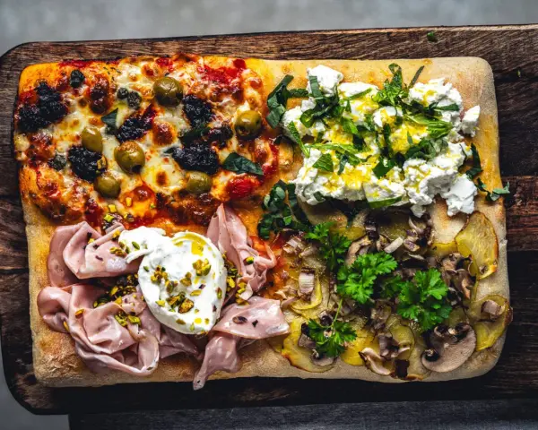 Vier verschiedene Beläge auf einer rechteckigen pizza al taglio: Oliven, Ricotta mit Kräutern, Mortadella mit Burrata und Kartoffeln mit Pilzen.