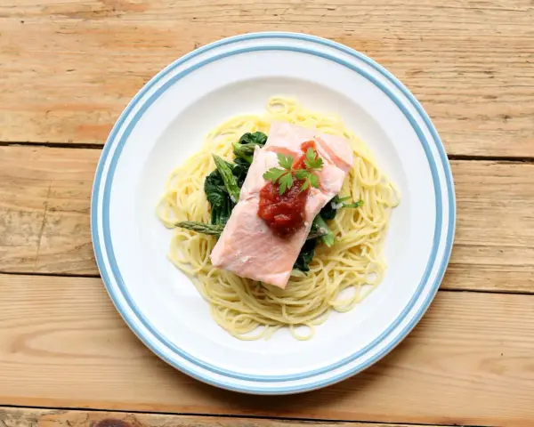 Ein Teller mit One Pot Lachs Pasta, garniert mit Petersilie und Tomatensauce, daneben Spargel und Spinat auf einem Holztisch.