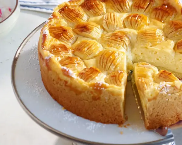 Ein köstlicher, schneller Apfelkuchen mit Schmand und Pudding, verziert mit Apfelscheiben und Puderzucker. Daneben eine Schüssel mit Beeren-Sahne.