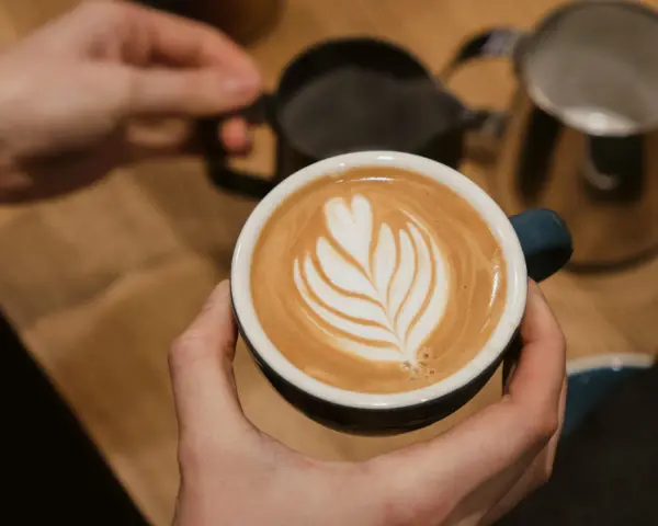 Cappuccino selber machen: Hände halten eine Tasse mit Latte Art. Im Hintergrund Milchkännchen und Sieb.