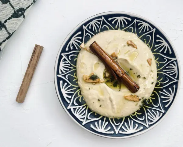 Cremiger Hummus, selbst gemacht, mit Zimtstange und Knoblauch garniert auf einem blau-weißen Teller.