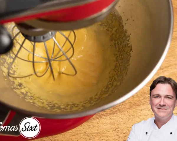 Küchenmaschine mixt eine cremige, gelbe Masse, die an Sauce Hollandaise erinnert. Ein Koch lächelt im Vordergrund.