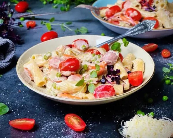 Cremige One Pot Pasta mit Würstchen, Erbsen und Kirschtomaten, garniert mit frischen Kräutern und geriebenem Käse.
