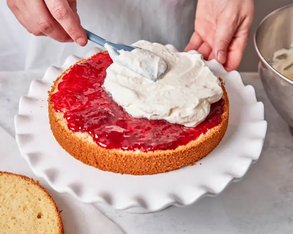 Eine Hand verteilt Sahne auf einer Marmeladenschicht eines Victoria Sponge Cake.