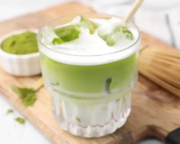Erfrischender Matcha Latte mit Eis, Milch und einem Hauch von Starbucks-Rezept-Magie. Daneben eine Schale Matcha-Pulver und ein Chasen.