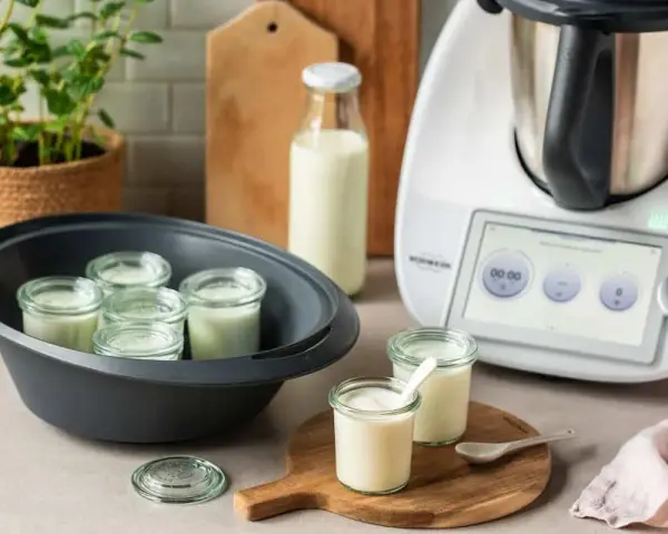 Kleine Gläser mit Joghurt in einem Dampfgarer, bereit, trinkjoghurt selber machen. Daneben ein Thermomix und Milchflasche.