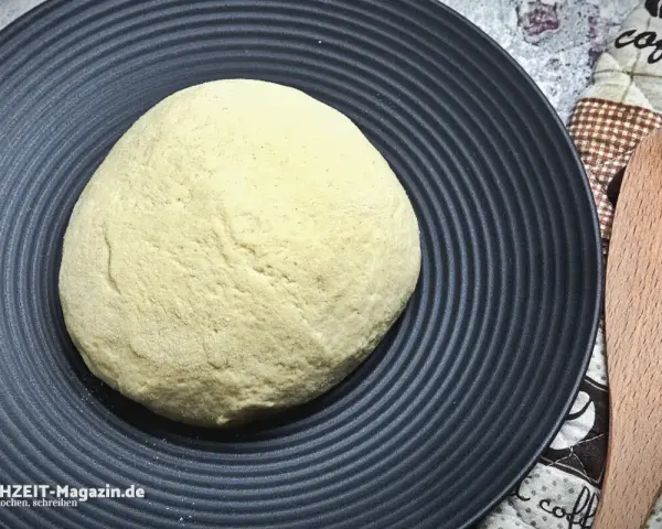 Ein runder Pizzateig auf schwarzer Platte wartet darauf, zu wissen, wie lange muss ein pizzateig ruhen, bevor er perfekt wird.