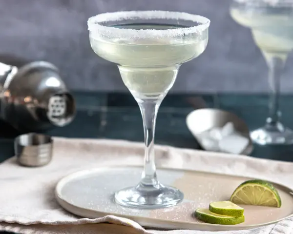 Ein perfekt zubereitetes Margarita-Rezept: Ein Glas mit Salzrand, daneben Limettenspalten, Eiswürfel und ein Shaker.