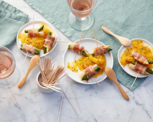 Leckere Spargel im Speckmantel auf gelbem Chutney, serviert auf kleinen Tellern mit Holzspießen und Roséwein.