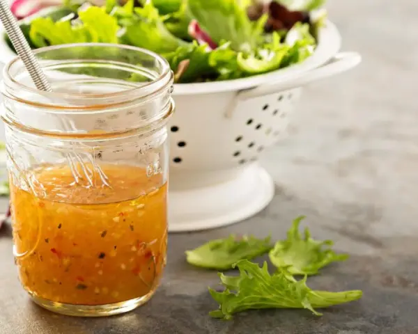 Ein Glas mit selbstgemachtem Salatdressing, das aussieht wie im Restaurant. Daneben ein Sieb mit frischem Salat.