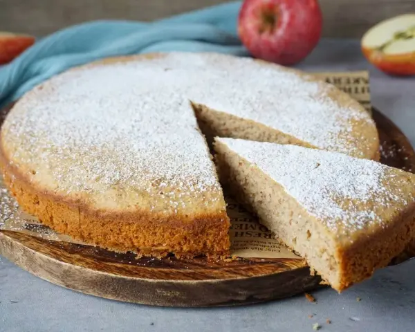 Ein schneller, saftiger Kuchen mit Apfelmus, bestäubt mit Puderzucker. Ein Stück wird gerade herausgenommen.