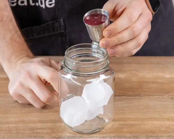 Eiswürfel in einem Glas, dazu wird rote Flüssigkeit aus einem Messbecher gegossen. So einfach lässt sich Rhabarberschorle selber machen!